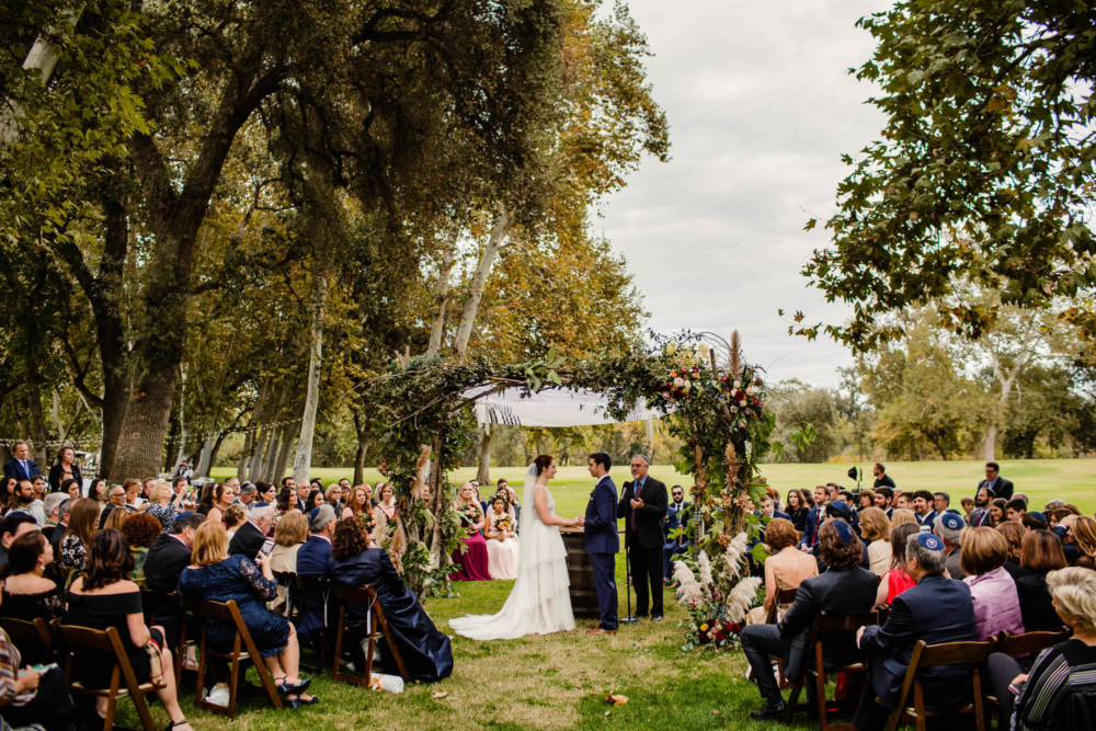 Guests, bride, and groom gathered for the wedding ceremony under large trees