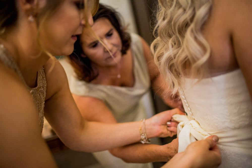 Bridesmaid and bride's mother help bride with her wedding dress