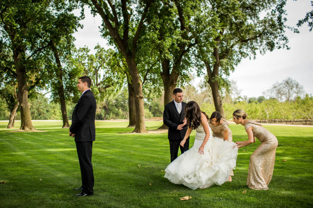 Bridesmaids adjusts bride's dress while groom waits with his back turned