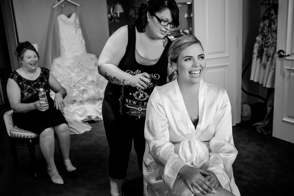 Bride smiles while having her hair done with her dress hanging in the background