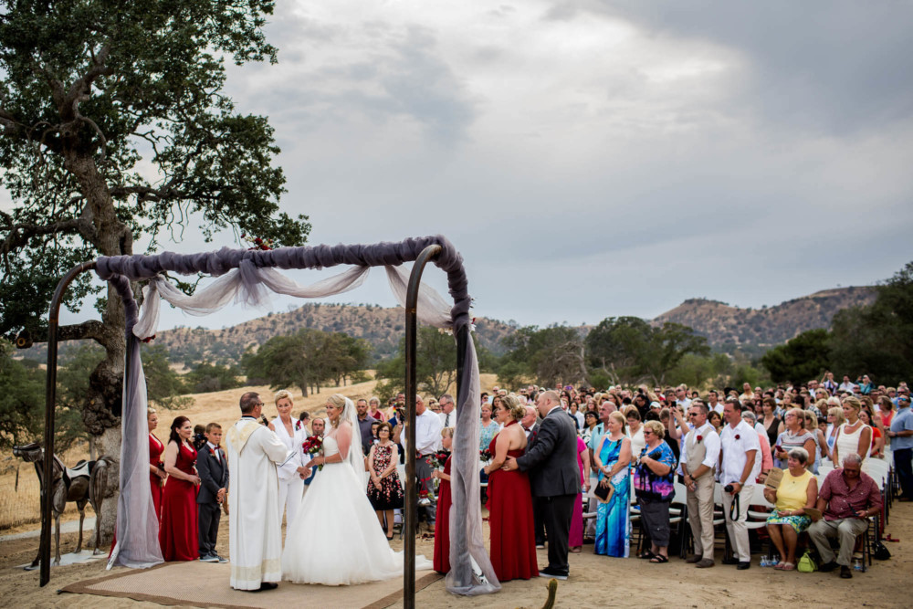 Brides and wedding guests during the ceremony under a large oak tree