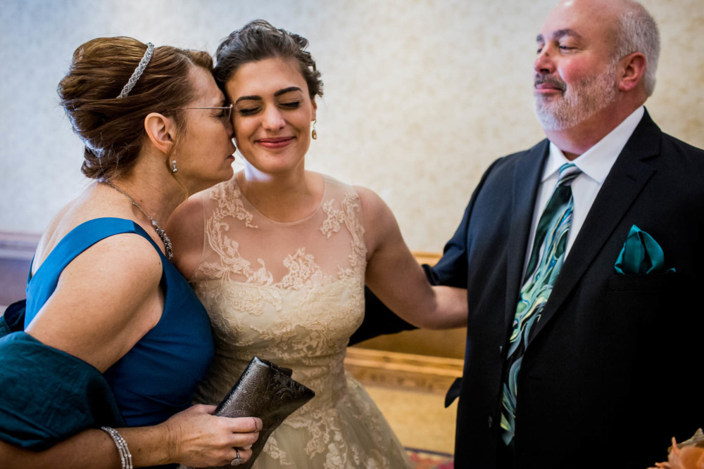 Bride's mother nuzzles her daughter while dad watches and smiles before the wedding