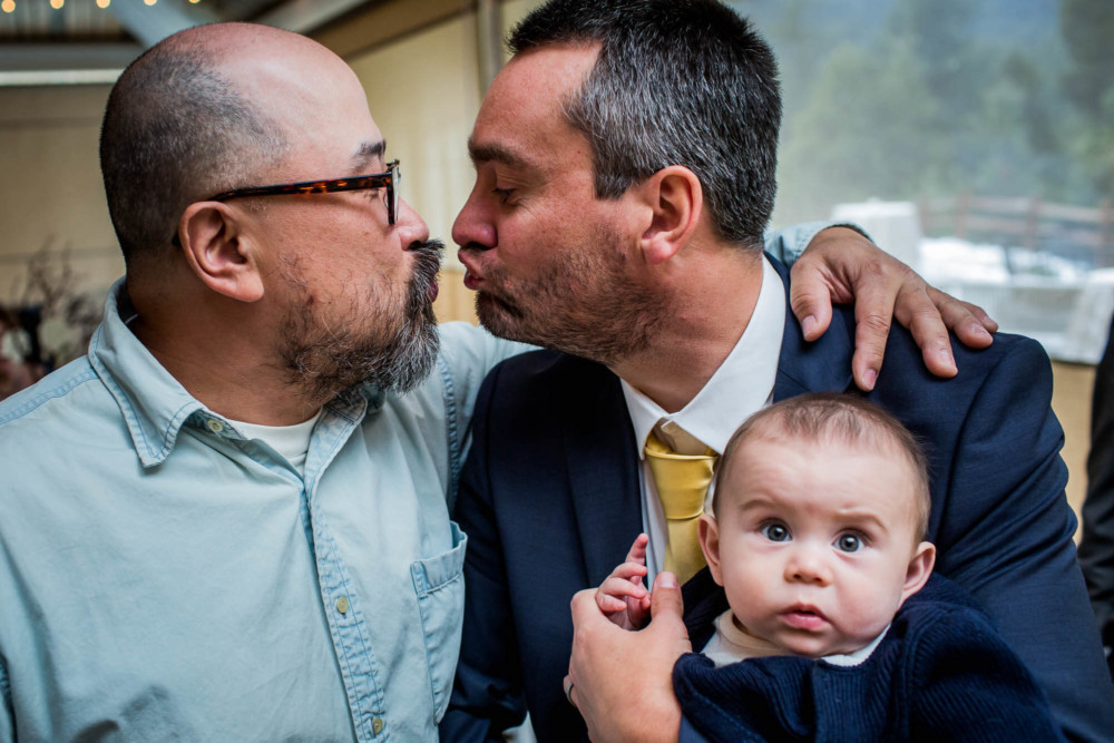 Groom and friend pretend to kiss while small baby makes a funny face