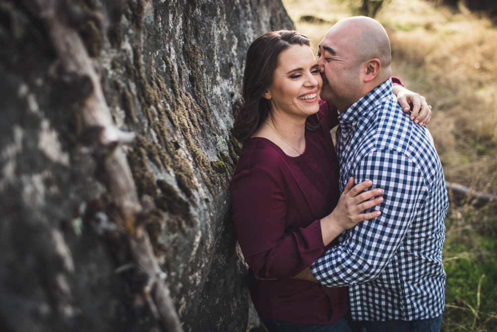 Groom kisses his bride as she leans on a rock during an engagement session