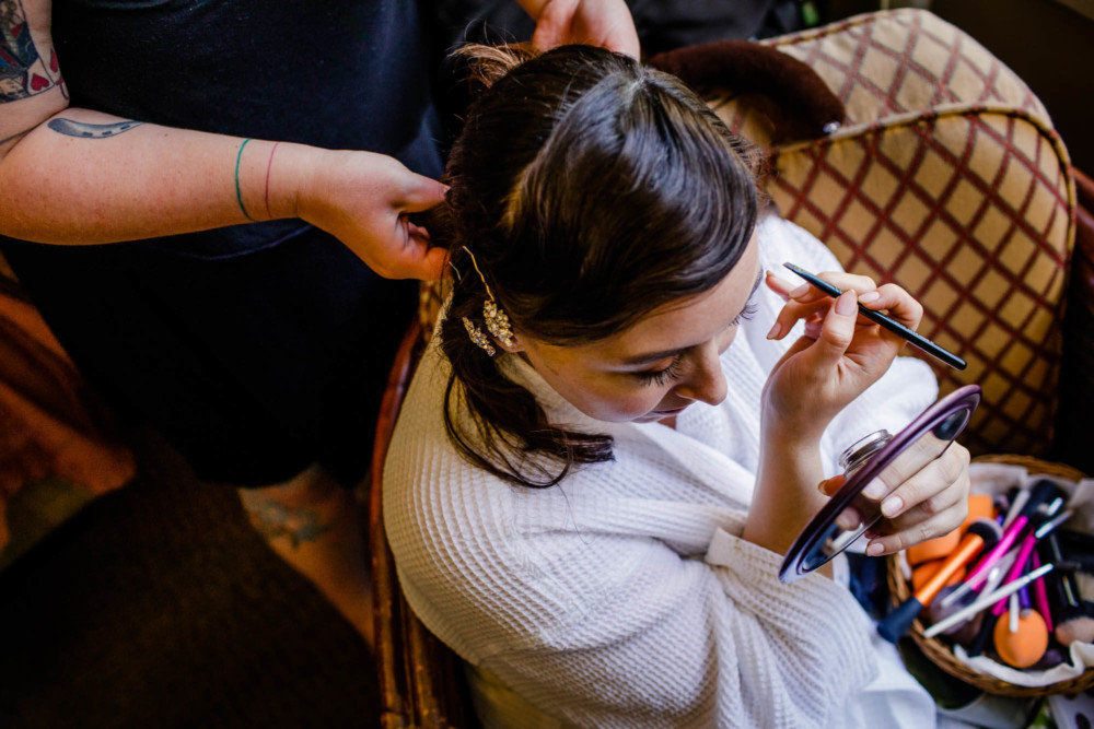 Bride works on her make up while a friend does her hair