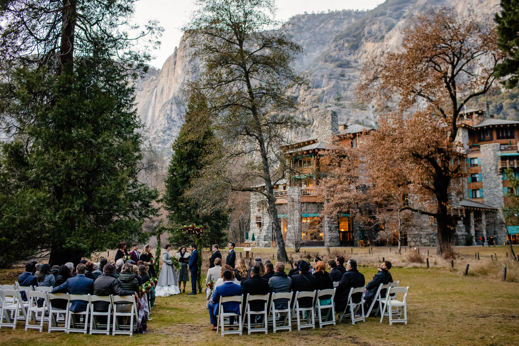 Wide shot of a wedding ceremony on the Majestic Yosemite Hotel wedding lawn with the hotel and Yosemite Falls in the background