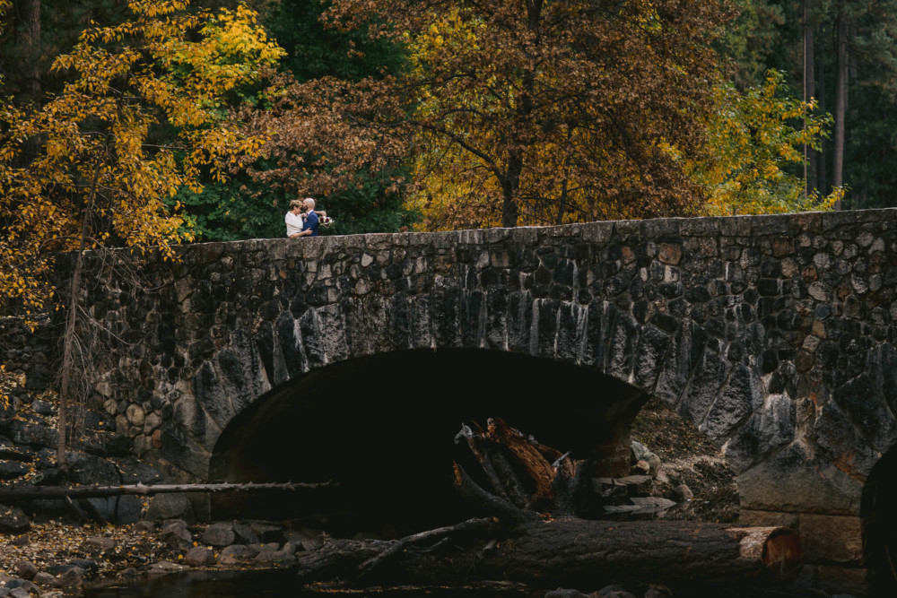 Bride and groom on a stone bridge over the Merced River with trees in full fall color