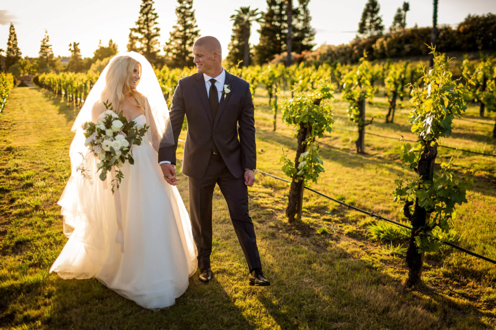 Bride and groom walk down the aisle of a vineyard at sunset