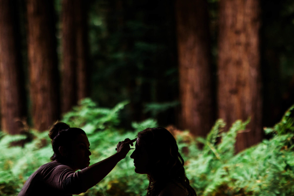 Silhouette of bride having her makeup done in front of ferns and redwoods