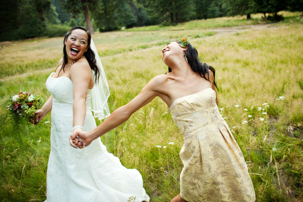 Bride and her bridesmaid hold hands and swing in a meadow.