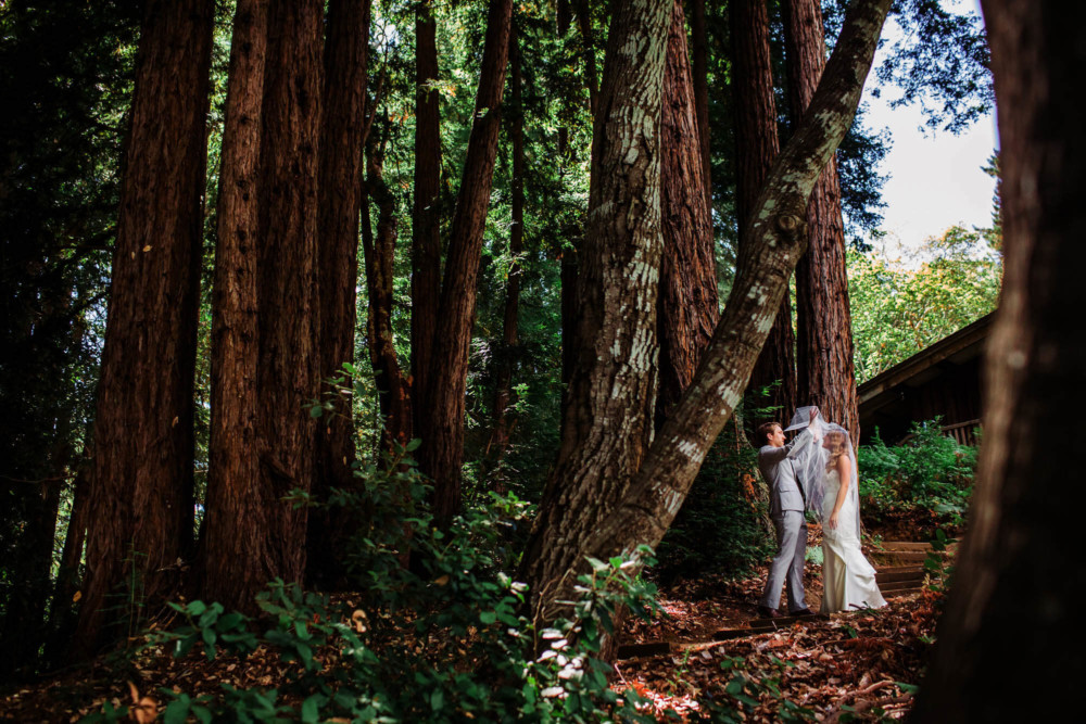 Groom lifts bride's veil during their first look among the redwoods at the Sequoia Retreat Center