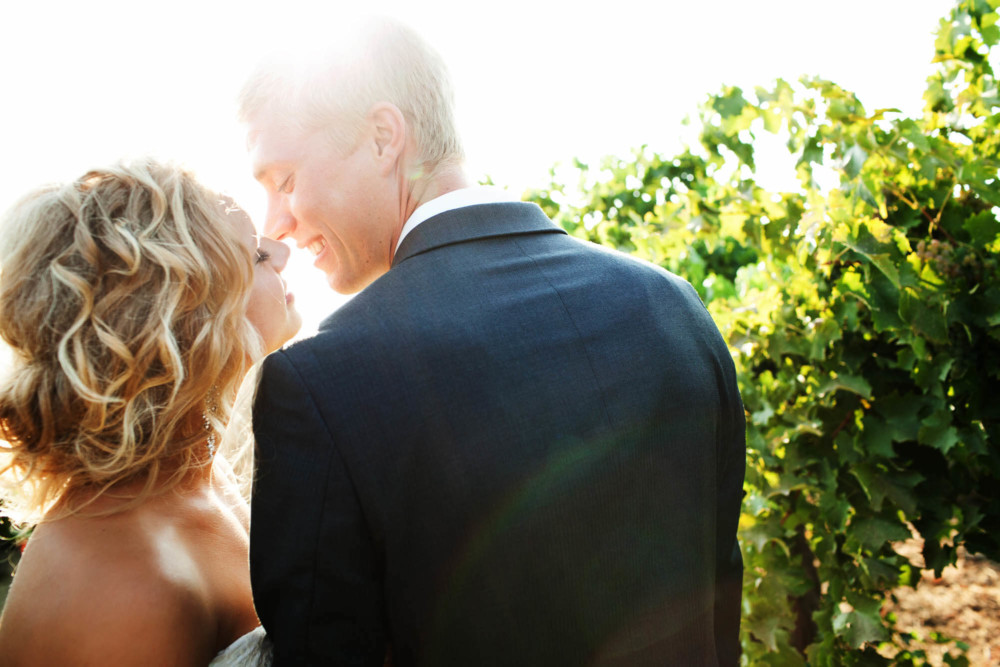 Bride and groom come close to kissing as the sun flares between them in a vineyard
