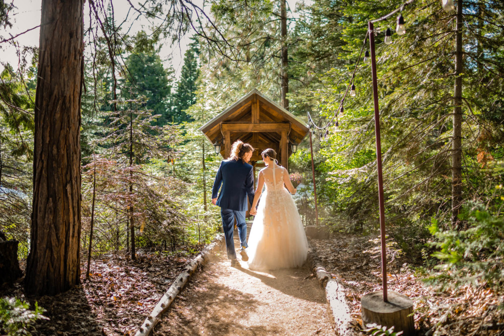Bride and groom walk hand in hand through the forest towards a covered stairway to their reception