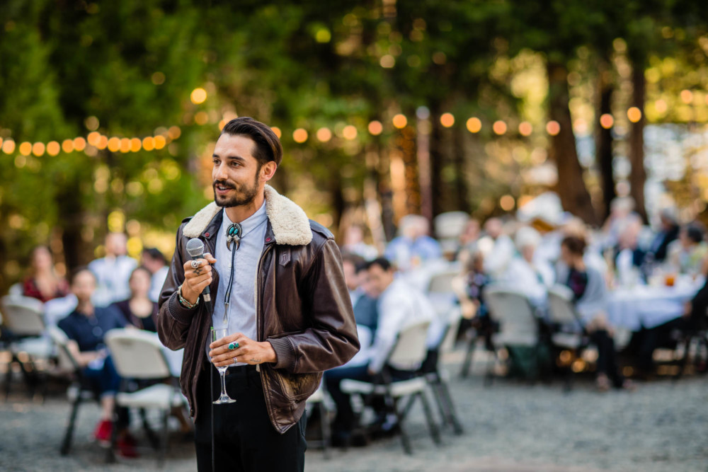 Man in a bomber jacket and bolo tie gives a speech at a wedding reception