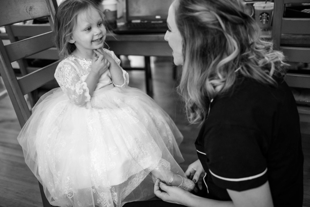 Flower girl smiles and laughs as a bridesmaid tries to put on her shoes