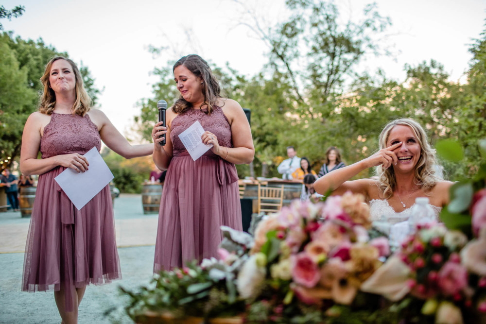 Maid of honor and bridesmaid laugh as bride wipes away a tear during the toasts at her wedding reception
