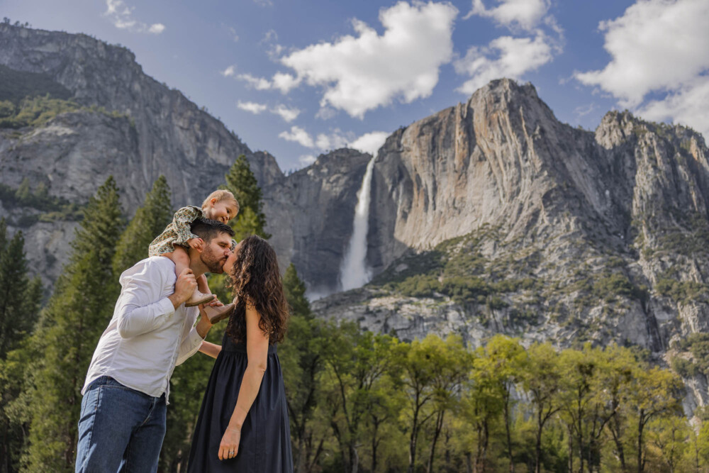 Mother and father kiss in front of a water fall while their daughter rides on his shoulders