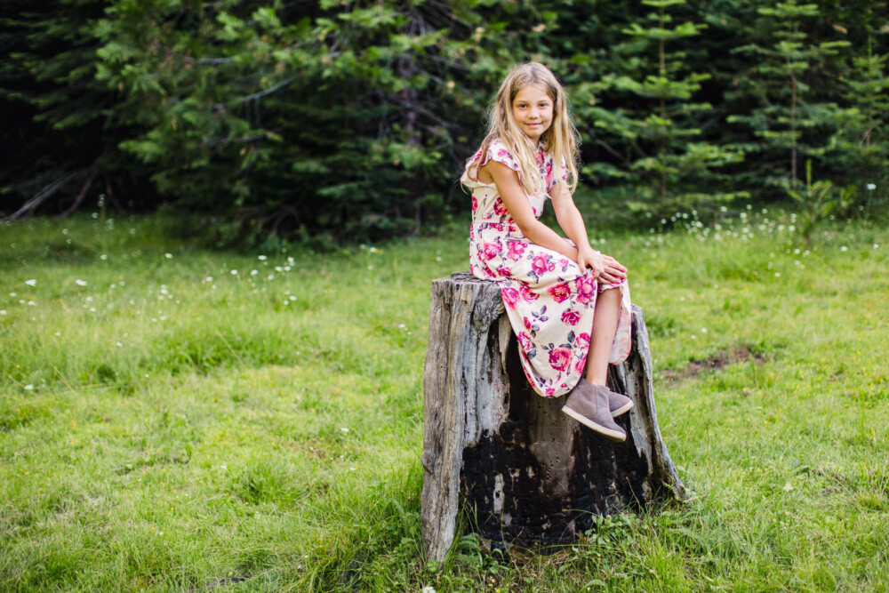 Girl in flowery long dress sits on old tree stump in mountain meadow