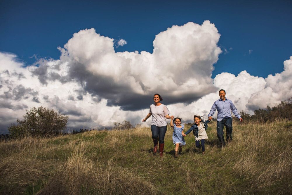 family of 4 runs down a hillside with big puffy white clouds in the sky