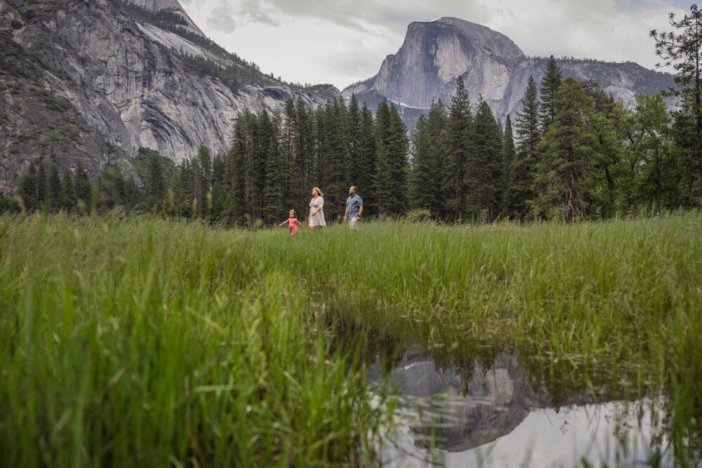 Family with a small child walks through Cooks meadow in Yosemite with Half Dome in the background and reflected in a pool of water