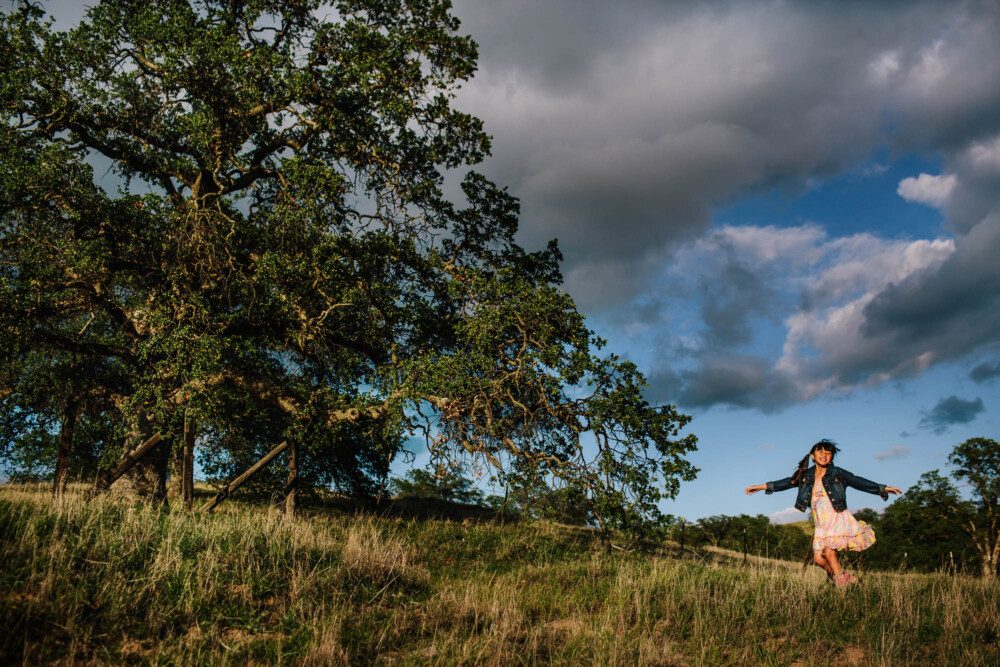 Young girl spins on a green hillside with large oak tree and dramatic clouds