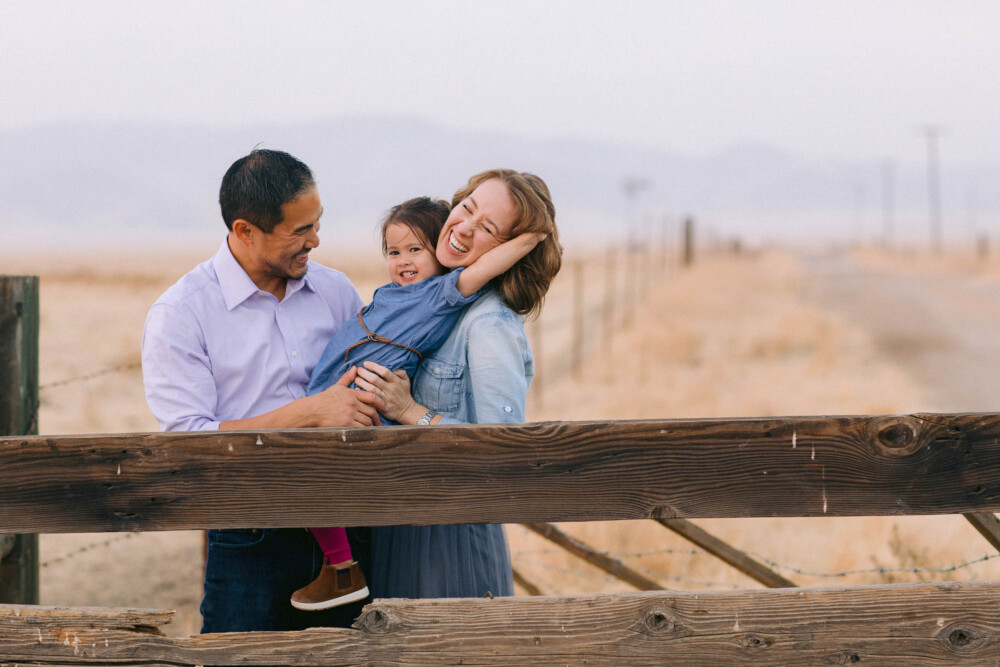 Multi-racial family cuddles and laughs on a ranch fence with foothills and windmill in the background