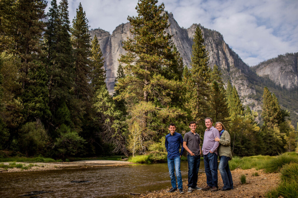 Family of four on a river bank in Yosemite with Yosemite Falls in the background