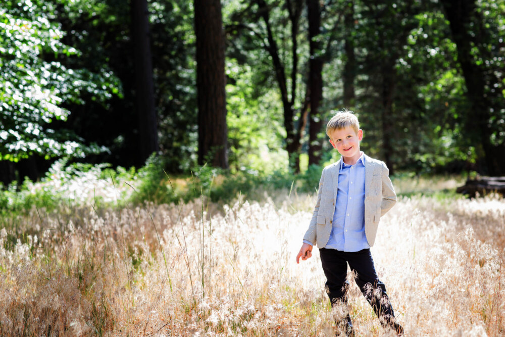 boy in a suit coat and whimsical look standing in a glowing golden meadow in the mountains