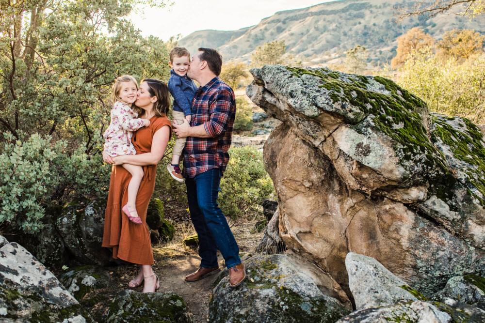 Mother and father kissing their children on a granite outcropping next to a large manzanita bush with Sierra foothills in the distance.